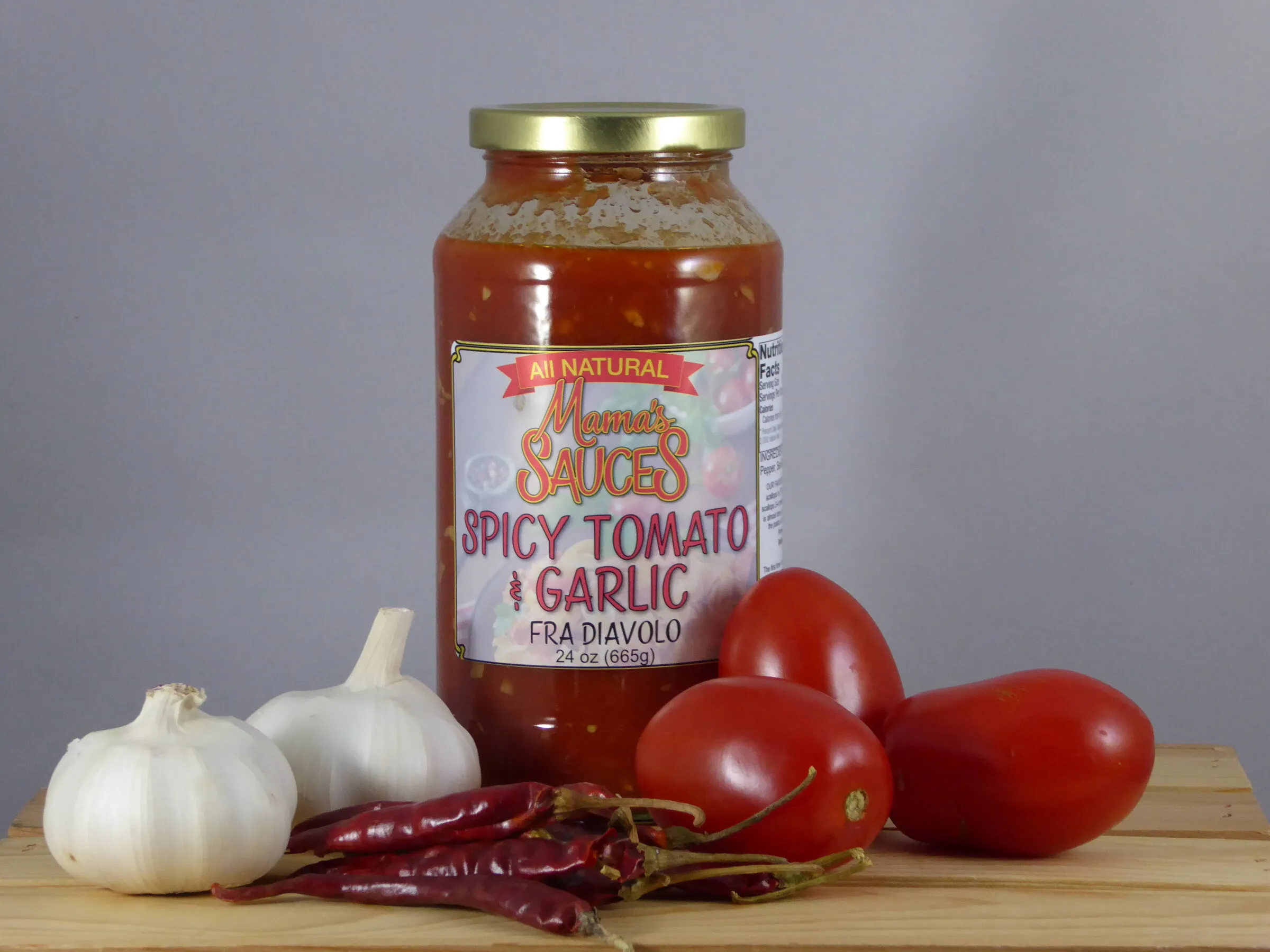 A jar of Mama's Fra Diavolo on a wooden crate with heads of garlic, plum tomatoes and dried red chilies